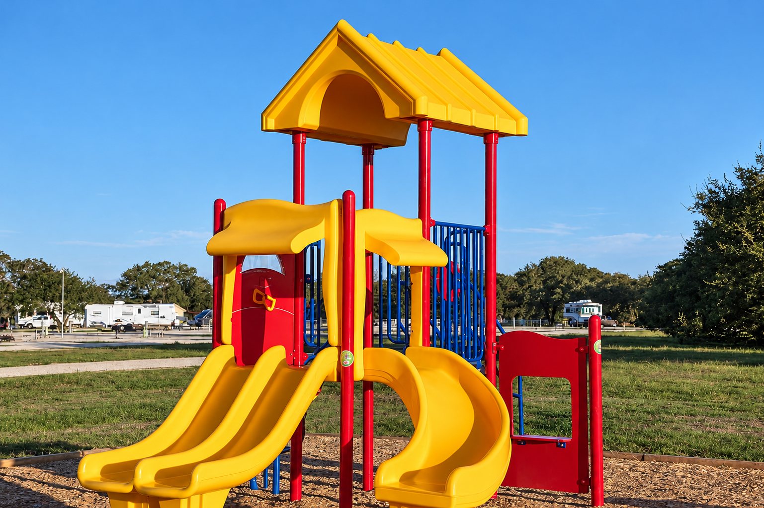 Une structure de jeux rouge et jaune avec deux toboggans jaunes, installée sur des copeaux de bois dans un parc herbeux sous un ciel bleu.