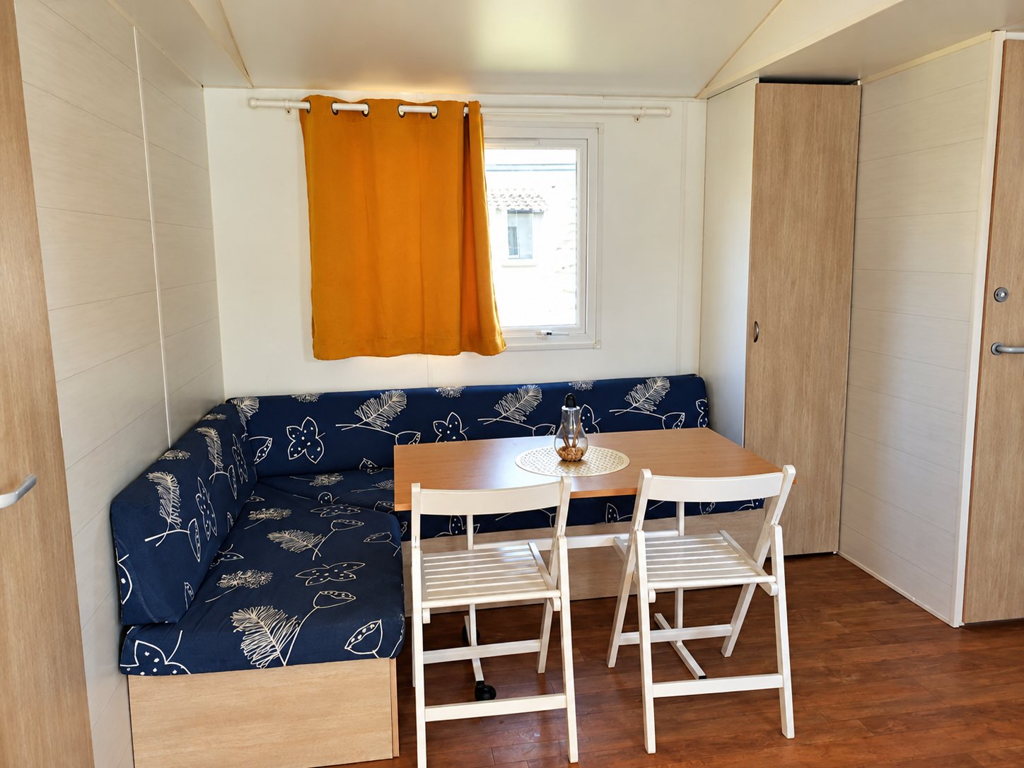 Intérieur d'un mobil-home avec une banquette d'angle à motifs bleus, une table en bois, deux chaises blanches pliantes et un rideau orange devant la fenêtre.