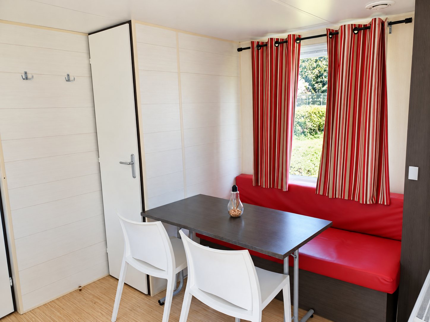 Intérieur d'un mobil-home avec une table sombre, deux chaises blanches, une banquette rouge et des rideaux rayés rouge et beige devant la fenêtre.
