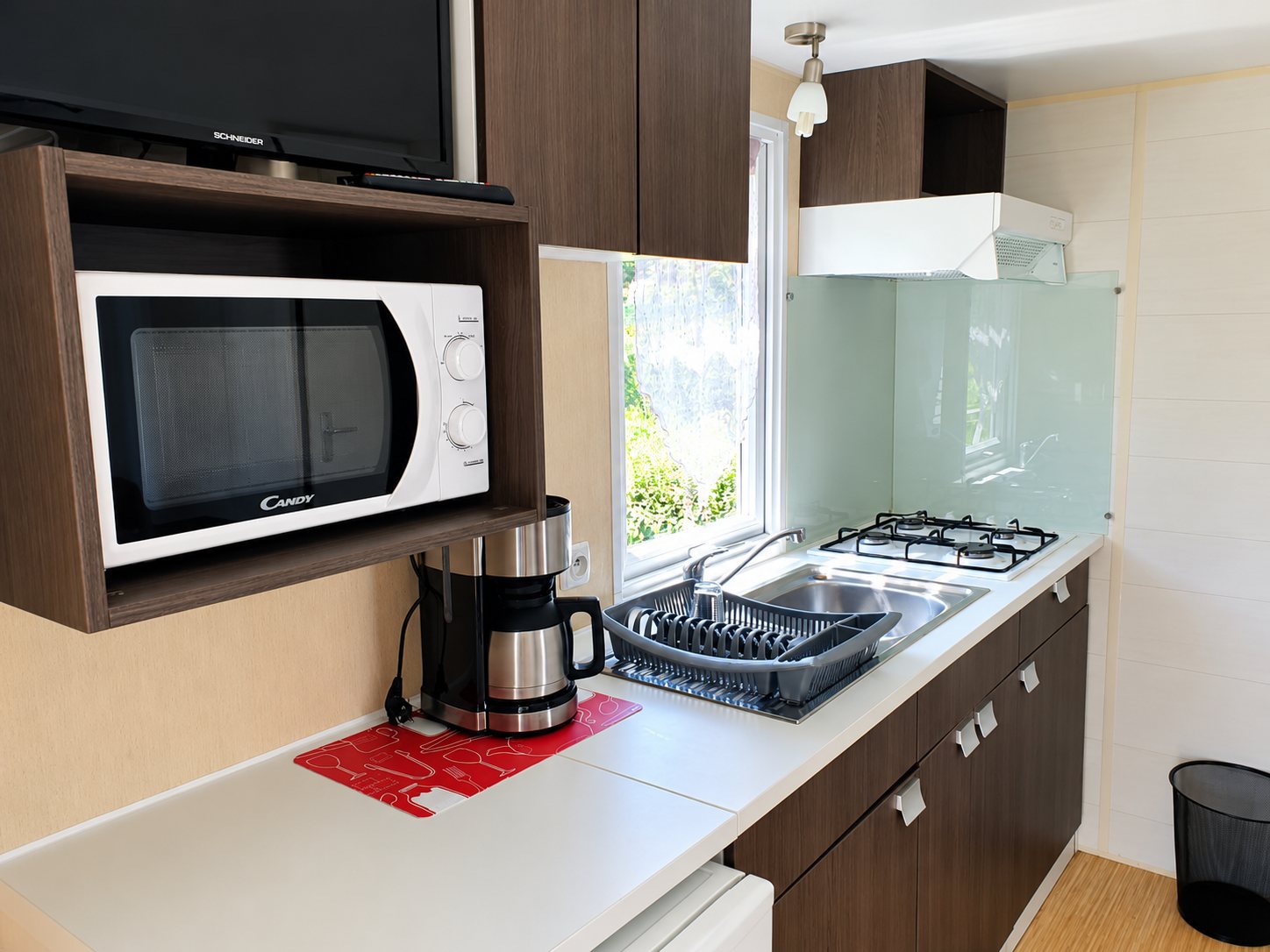 Coin cuisine d'un mobil-home avec un four à micro-ondes sous un téléviseur, une cafetière, un évier et une plaque de cuisson à gaz.