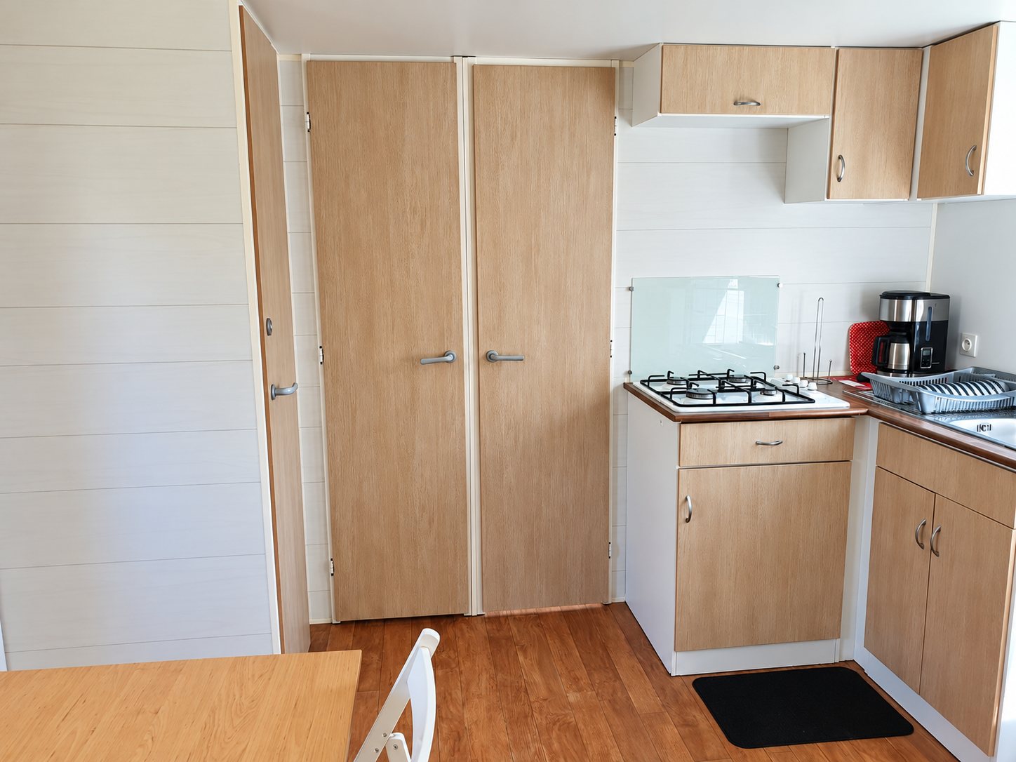 Intérieur d'un mobil-home avec une kitchenette équipée d'une plaque de cuisson à gaz, d'une cafetière et de placards en bois clair, à côté de deux grandes portes de rangement.